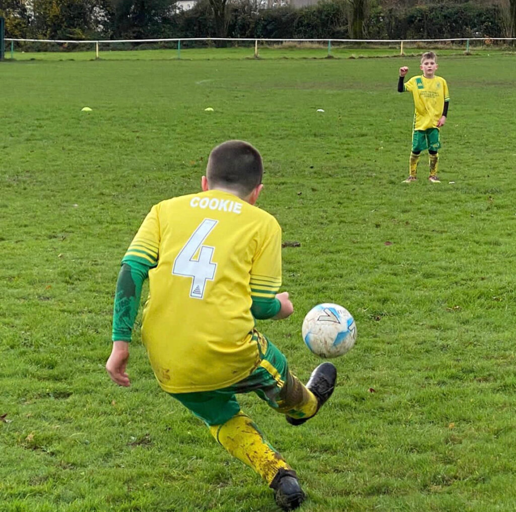 A boy in a Pendle football kit kicks a football across the pitch