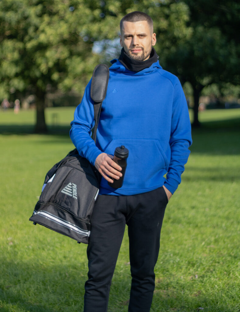 Model wearing a royal blue hoodie, black joggers and a black snood from the Pendle Lifestyle collection. He's also carrying a Pendle kit and water bottle. 