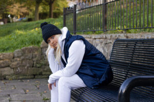 Female mode; wearing a black bobble hat, a navy gilet, and light grey hoodie and jogging bottoms,.