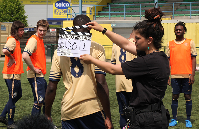 A film set with footballers in Pendle football kit waiting for the shoot to begin. A woman holds a clapperboard.